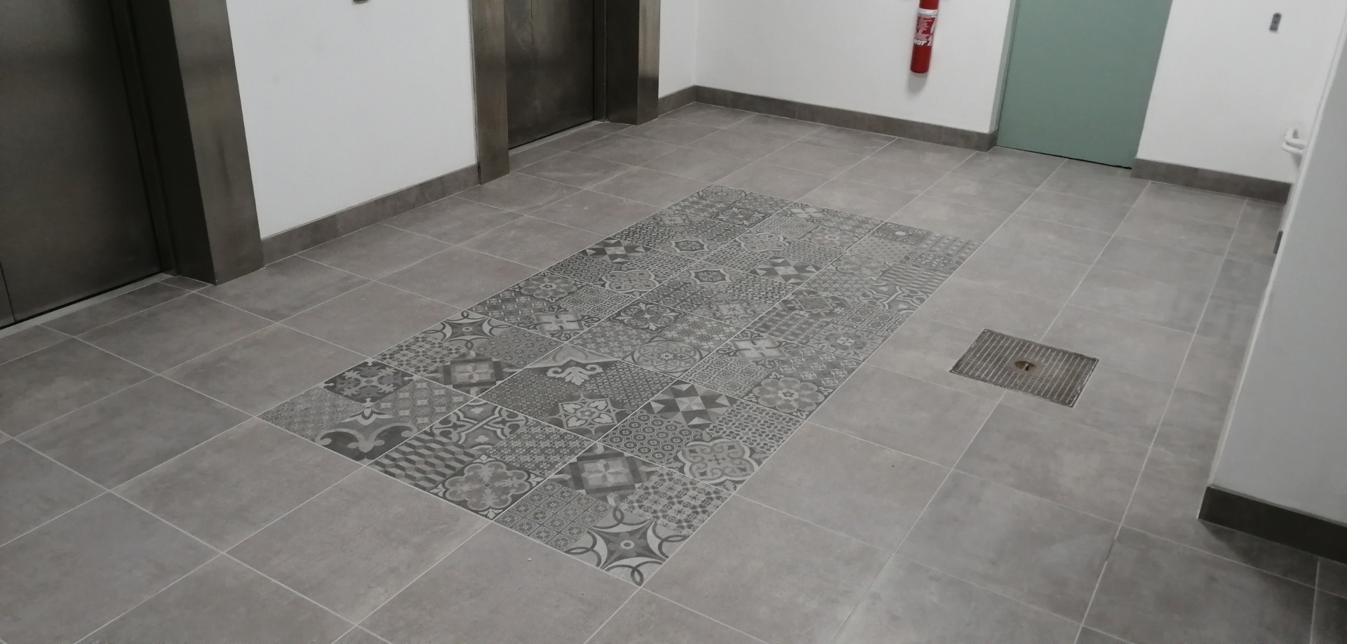 Sol en carrelage gris avec motif décoratif central en carreaux de ciment noirs et blancs dans un couloir moderne, équipé d'un extincteur mural et de portes en métal.