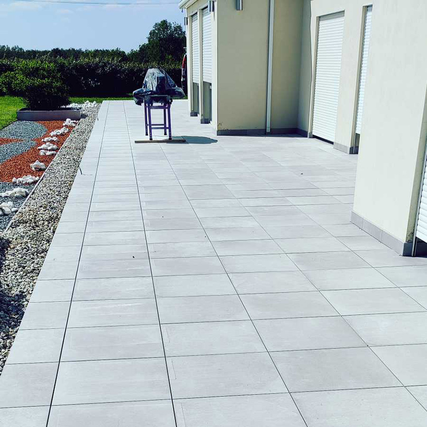 Terrasse extérieure carrelée en dalles grises, bordée de galets décoratifs et d'un espace vert, avec un barbecue recouvert en arrière-plan.