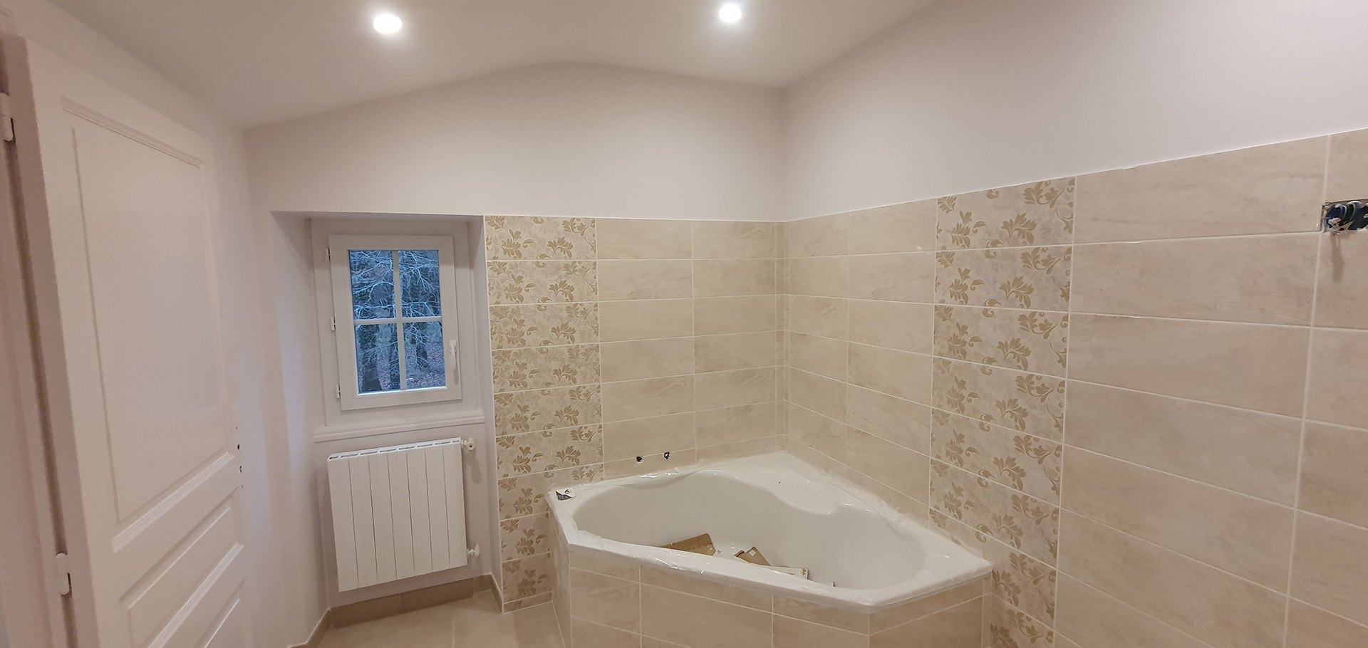 Salle de bain moderne avec baignoire d'angle, carrelage mural décoré de motifs floraux beige, radiateur vertical et fenêtre donnant sur l'extérieur.