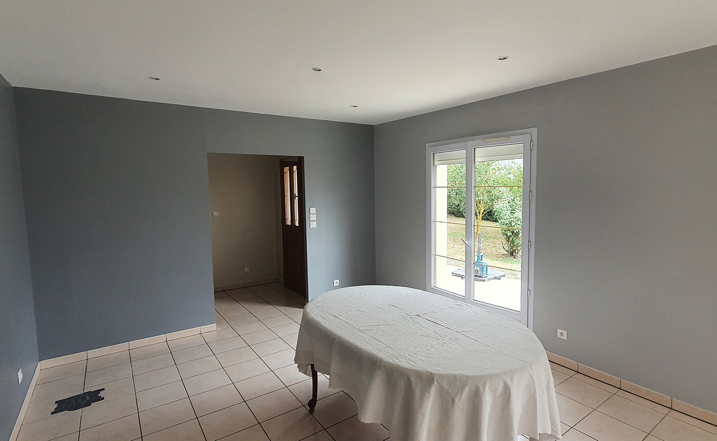 Pièce vide avec murs peints en gris clair, sol carrelé beige, une table ronde recouverte d'une nappe blanche, et une porte-fenêtre donnant sur un extérieur verdoyant.
