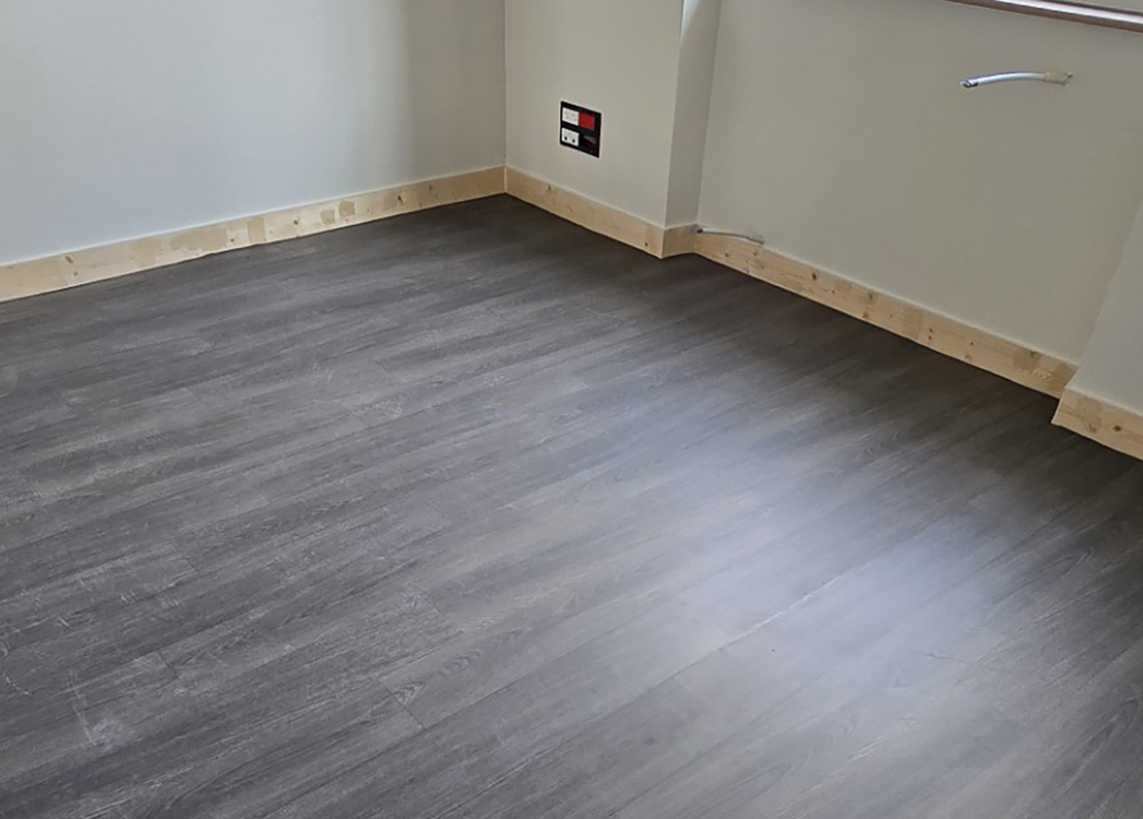 Sol stratifié gris foncé nouvellement posé dans une pièce, avec plinthes en bois clair et prises électriques murales visibles.