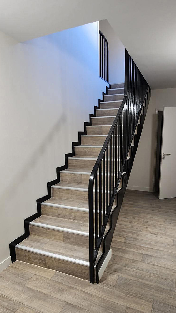 Escalier intérieur moderne avec marches en stratifié imitation bois, contremarches noires et rampe métallique noire, menant à un étage supérieur éclairé par une fenêtre.